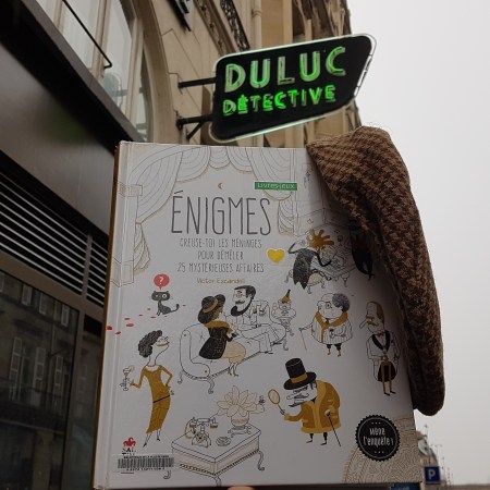 Photographie du livre "Enigmes, creuse toi les méninges pour démêler 25 mystérieuses affaires, de Victor Escandell, avec une casquette de détective devant une agence parisienne de détectives pour le blog littéraire Mangeurdelivres