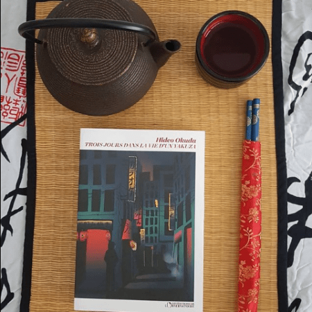 Photo du livre sur une natte en bambou avec une théière japonaise et une tasse de thé ainsi qu'une paire de baguettes.