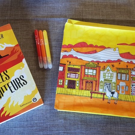 Photo du livre à côté d'un coloriage d'un décor de Western dans les trois 4 teintes utilisées par la couverture du livre : jaune, orange, rouge et blanc